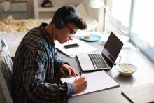 Teenage boy wearing headphones works at desk in his bedroom.